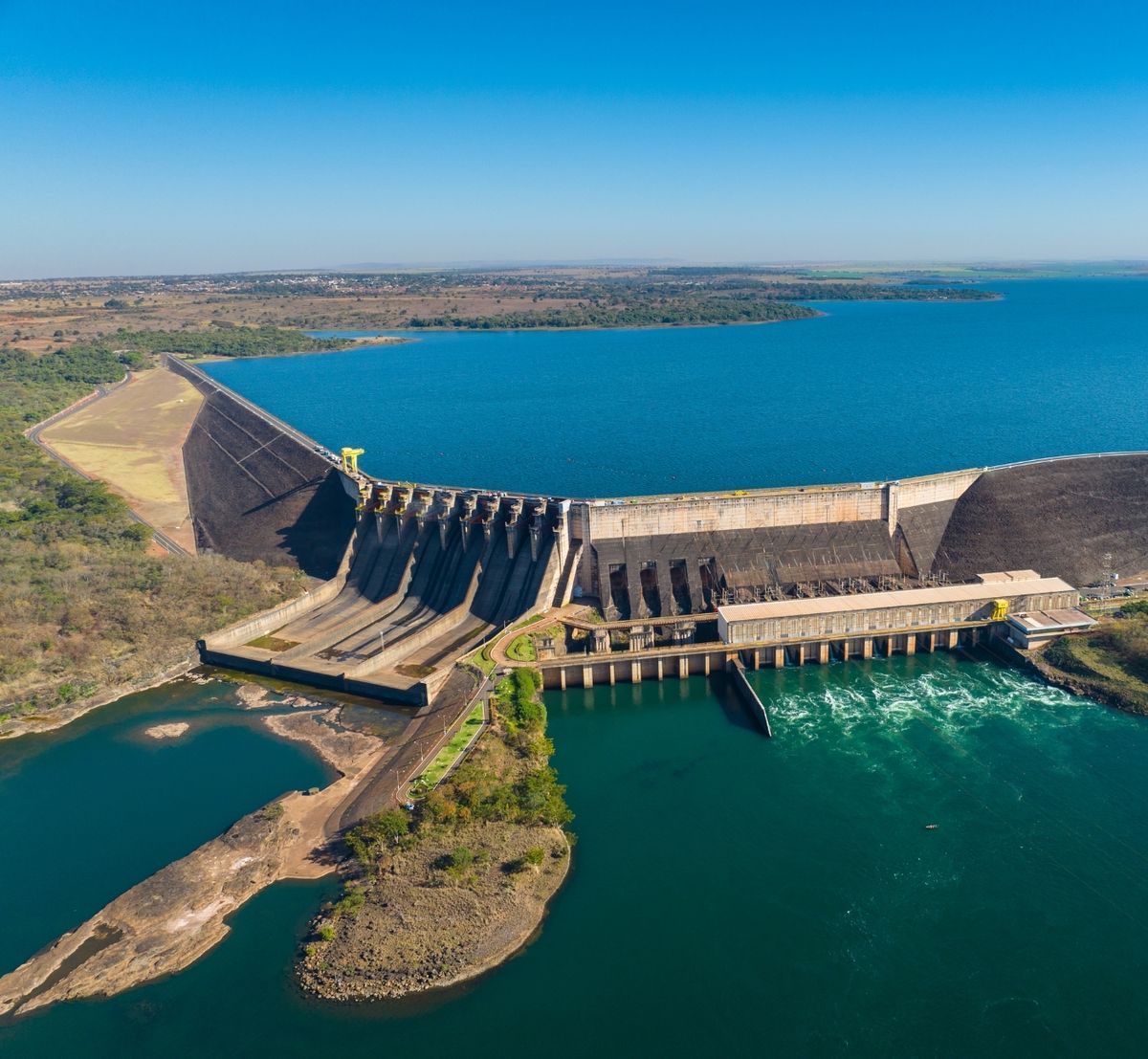 Vista aérea de uma barragem de concreto com vertedouro e casa de força, entre um reservatório e o rio.