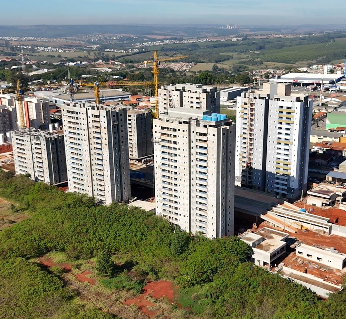 Vista aérea de edifícios residenciais altos em construção, com gruas amarelas e área verde em primeiro plano.