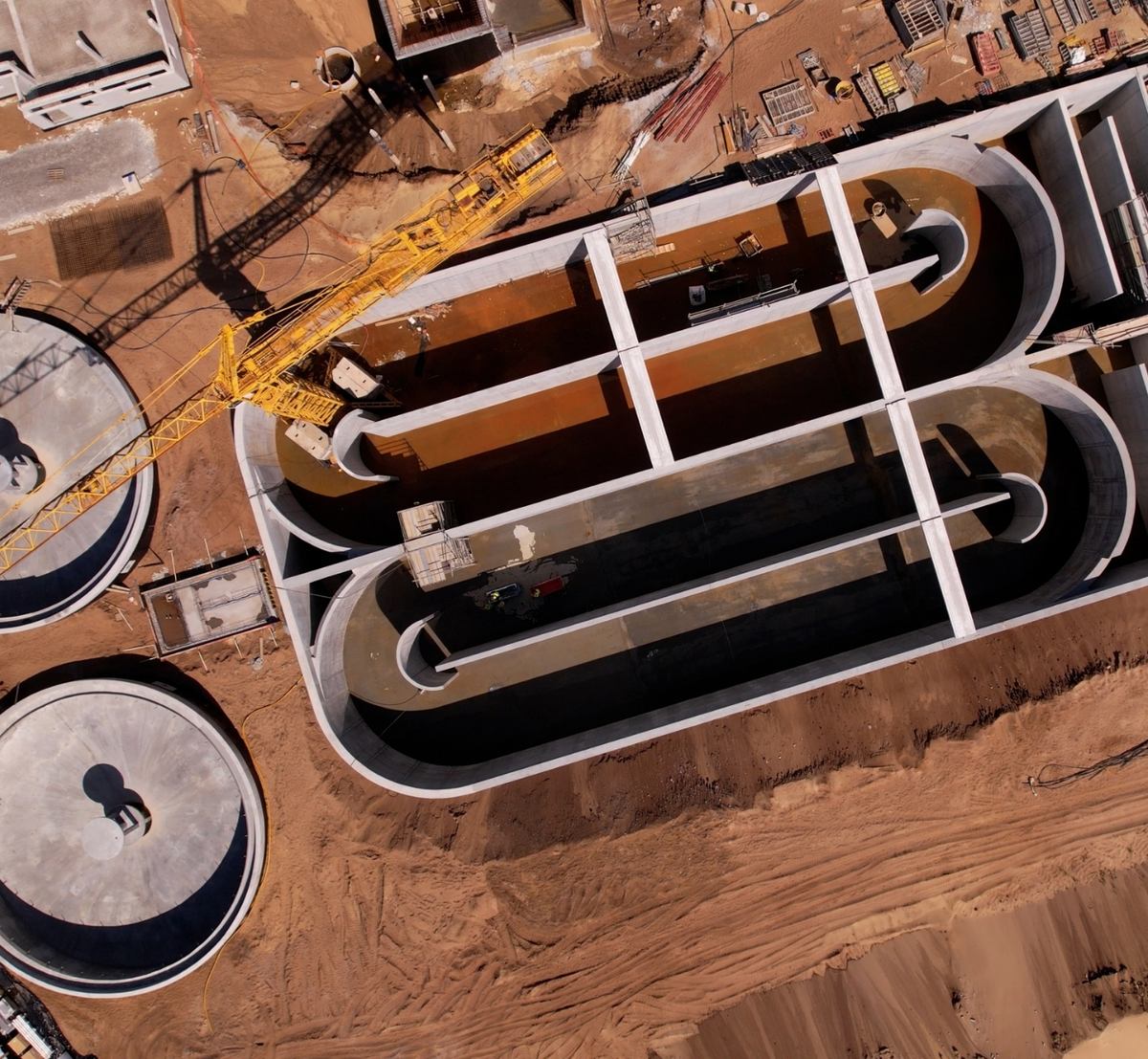 Vista aérea vertical de tanques de concreto em construção, com divisórias curvas, decantadores circulares e uma grua amarela.